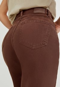 Jean en denim marron taille haute avec une silhouette ajustée, doté d'une poche arrière et d'une texture côtelée subtile. Étiquette de marque en cuir sur la ceinture.