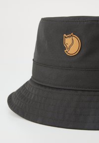 Chapeau seau noir en tissu, doté d'un bandeau cousu et d'un logo de renard en relief couleur sable à l'avant. Surface texturée avec des stries.