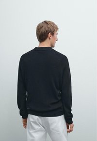Zwarte gebreide pullover met een kraag, lange mouwen en een geribbelde textuur. De achteraanzicht toont een relaxte pasvorm en een eenvoudige uitstraling.