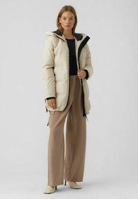 Beige gewatteerde jas met capuchon, voorzien van een zwarte voering en ritssluiting. Draagt over een zwarte top, in combinatie met lichtbruine wijde broek.