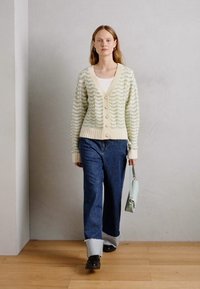 Cardigan verde-claro com padrão ondulado texturizado, com acabamentos em creme e botões de pérola. Combinado com jeans de perna larga e uma bolsa azul-claro.