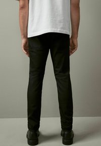 Mustat slim-fit-housut, joissa on sileä pinta, varustettuna takataskuilla, yhdistettynä mustiin kenkiin. Taustana on neutraali harmaa.