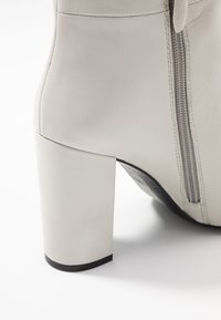 Botín de cuero beige hasta el tobillo con punta cuadrada, un tacón bloque robusto y cremallera lateral. Textura suave con un brillo sutil.