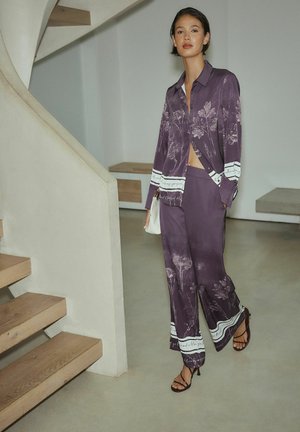 Lila Hemd mit Blumenmuster und passende weite Hosen aus seidigem Material mit weißen Akzenten, ergänzt durch Riemchenabsätze.