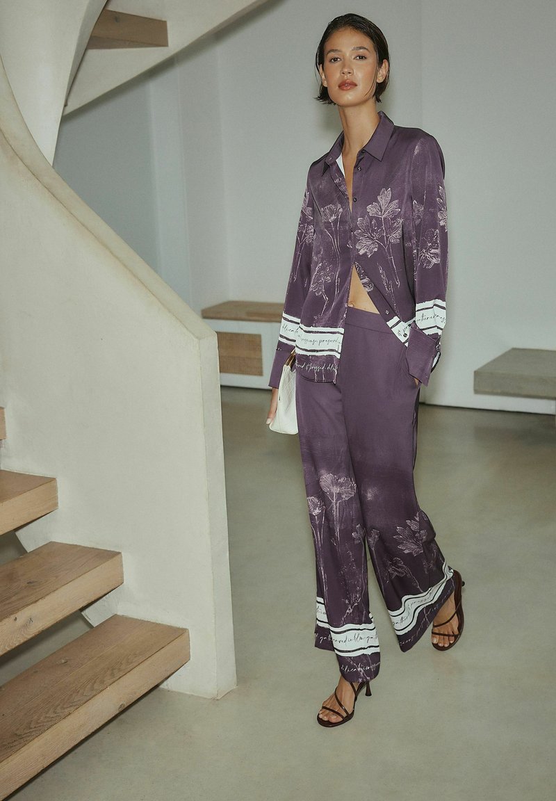 Lilla skjorte med blomsterprint og matchende vidde bukser, lavet af silkeagtigt materiale med hvide detaljer, suppleret med stropper på hælene.