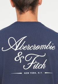 Námořnická modrá tričko s bílým kurzívním textem "Abercrombie & Fitch" a "NEW YORK, N.Y." na zadní straně, vyrobené z měkké tkaniny.