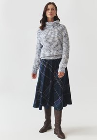 Pull en tricot dans des tons de bleu clair avec un col haut, associé à une jupe carrée bleu marine avec des lignes vertes et violettes. Bottes marron hauteur genou.