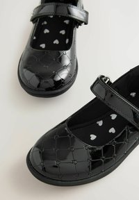 Svarta lackskinnsskor med Mary Jane-design, dekorerade med hjärtmönster och en kardborreband. Insidan har ett prickigt foder med små hjärtan.