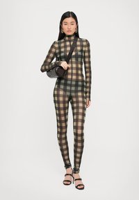 Black and olive plaid bodysuit with a high neck, sheer texture, long sleeves, and fitted legs, paired with a small black crossbody bag.
