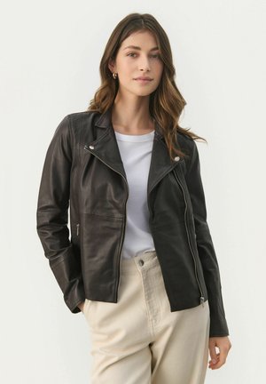 Black leather motorcycle jacket with a zip closure, asymmetrical collar, and silver hardware accents. Worn over a white shirt and beige pants.