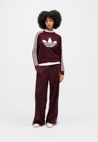 Burgundský pletený sveter s bielym logom adidas, v kombinácii so zodpovedajúcimi širokými nohavicami so tromi bielymi pruhmi po stranách a jemnou textúrou.