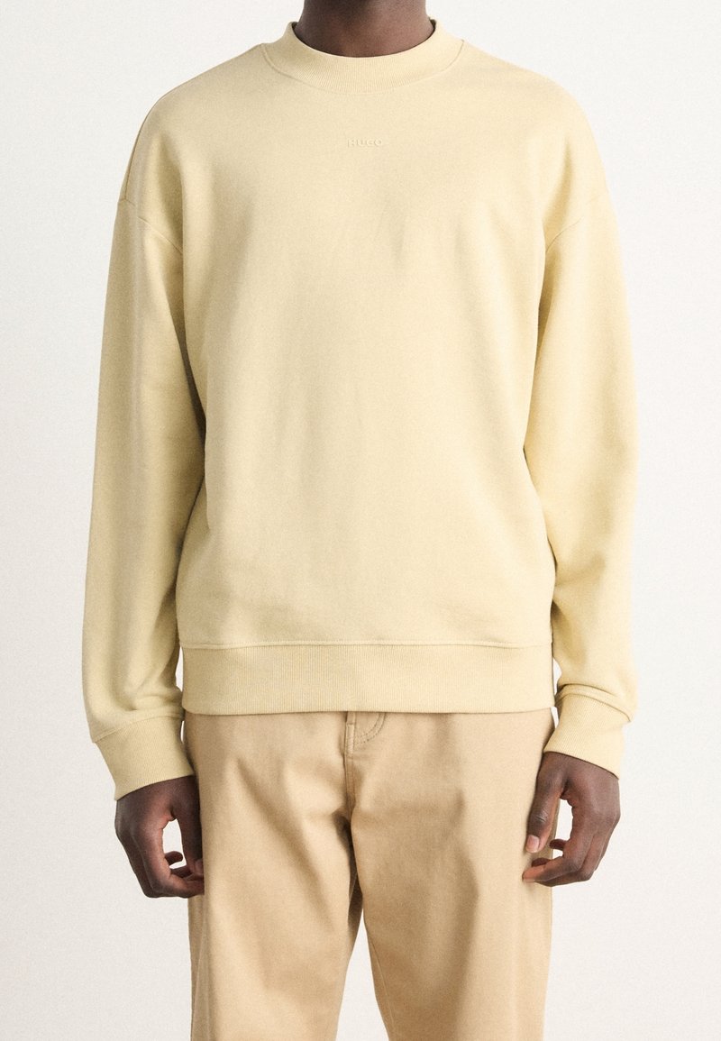 Personne portant un sweat-shirt beige clair à manches longues et un pantalon beige assorti, debout devant un fond blanc uni.