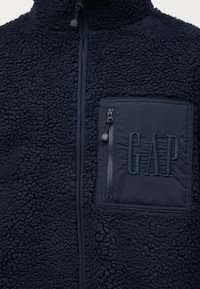 Μπουφάν από φλις σε ναυτικό μπλε χρώμα με ψηλό γιακά, κεντρικό φερμουάρ και τσέπη στο στήθος με φερμουάρ, που φέρει το κεντημένο λογότυπο "GAP".