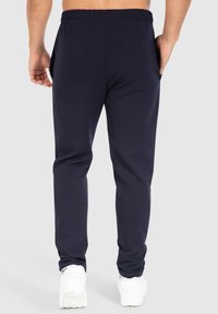 Hombre con joggers ajustados en color navy y cinturilla elástica, usando zapatillas blancas, de pie con las manos relajadas a los lados, vista desde atrás.
