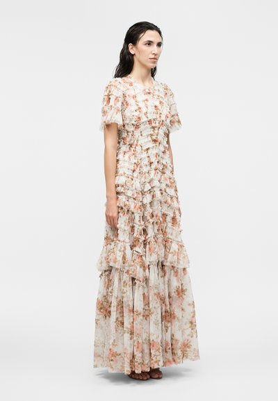 Γυναίκα φορώντας ένα μακρύ, ανθισμένο φόρεμα με βολάν, κοντά μανίκια και φούστα με στρώσεις, στέκεται μπροστά σε απλό λευκό φόντο.