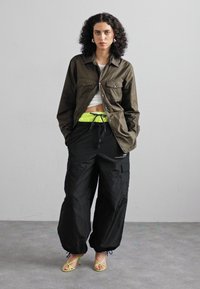 Olijfgroene button-up jas met twee borstzakken, gecombineerd met zwarte cargobroeken met een drawstring taille en neongele accenten. Gele sandalen.