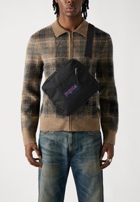 Juodas nailono per petį nešiojamas krepšys su priekiniais užtrauktuko kišenėmis, su mėlyna ir raudona "Jansport" logotipo etikete. Dėvimas ant rudo pašiltinto megztinio su languotais raštais.