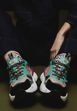 Persona con zapatillas gruesas y coloridas con acentos en teal, negro, blanco y estampado animal, cordones con patrón en blanco y negro, y detalles morados en el talón.