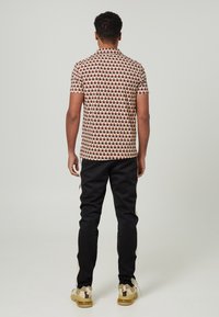 Chemise à manches courtes avec un motif géométrique rouge et beige, associée à un pantalon noir avec des bandes latérales et des baskets beiges à semelle texturée.
