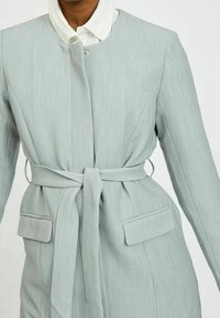 Cappotto su misura grigio chiaro con una texture liscia, dotato di vita fasciante, tasche laterali e colletto rotondo con chiusura nascosta.