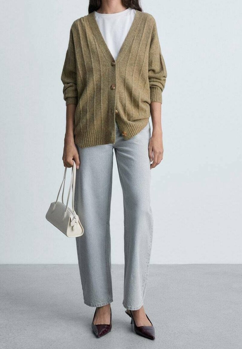 Personne portant un cardigan beige, une chemise blanche, un jean gris clair et des chaussures pointues foncées, tenant un petit sac à main blanc à ses côtés.