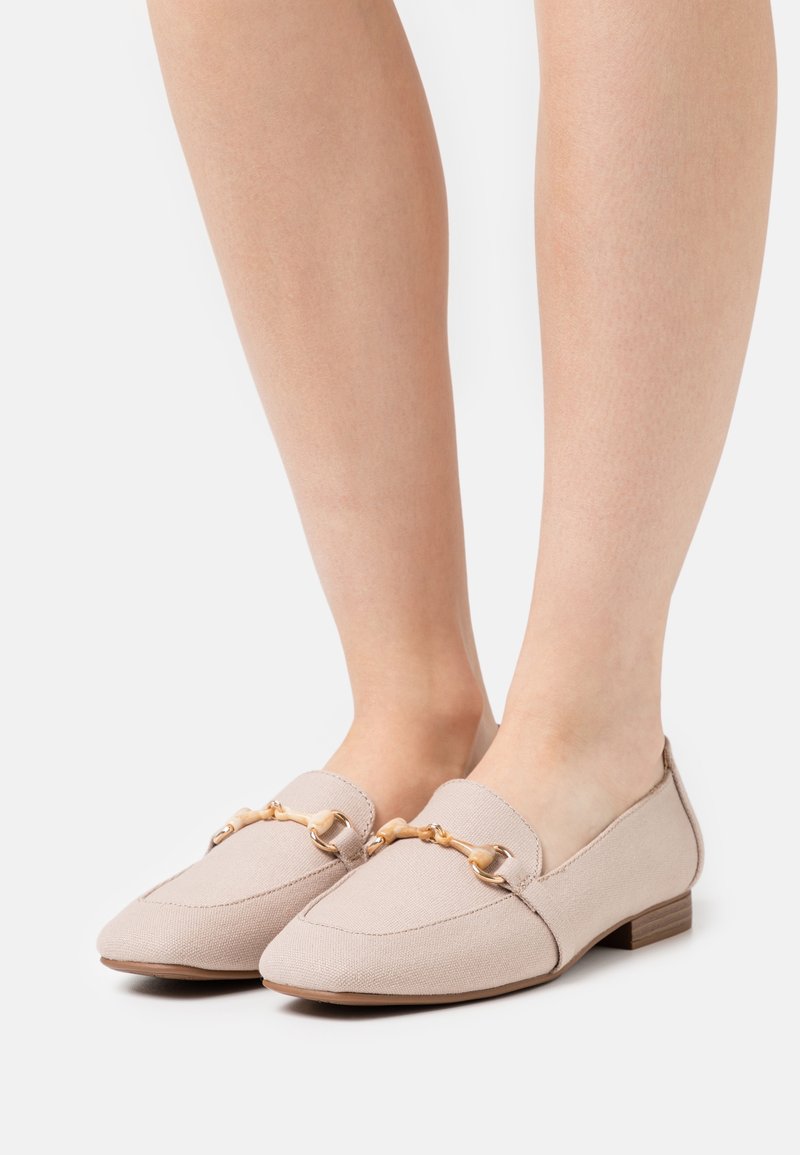 Mocassins en tissu beige avec détail mors en ton or portés aux pieds, dotés de bas talons empilés en bois et d'orteils pointus.