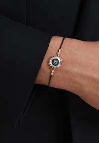 Guldtonat armband med en blå och svart cirkulär detalj, som har en läderrem och en stilren, minimalistisk design.