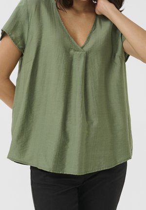 Femme portant un haut vert olive ample à manches courtes et col en V ainsi qu'un pantalon noir, une main reposant près de son cou.