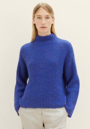 Woman wearing a blue textured knit sweater with a high collar and beige pants, standing against a light neutral background.