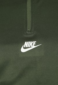 Felpa sportiva verde con zip, realizzata in un tessuto testurizzato. Presenta una zip parziale sul davanti e un logo Nike bianco nella zona sinistra del petto.