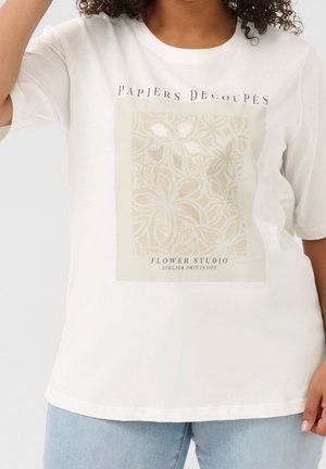 Kobieta nosząca białą koszulkę z beżowym kwiatowym wzorem i tekstem "Papiers Decoupés Flower Studio Atelier Printemps", połączoną z jasnoniebieskimi dżinsami.