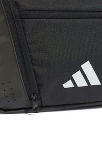Svart treningsbag med teksturert stoff, glidelåslomme og hvit merkevarelogo. Har ventilasjonsåpninger på siden.