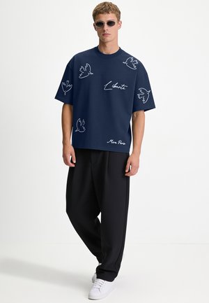 Giovane uomo con occhiali da sole ovali, t-shirt blu navy con disegni bianchi di uccelli e testo, pantaloni neri larghi e sneakers bianche su sfondo semplice.
