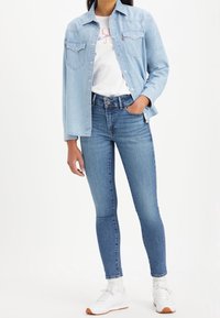 Lichtblauwe denim overhemd met twee borstzakken over een witte graphic t-shirt, gecombineerd met donkerblauwe skinny jeans en witte sneakers.