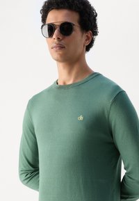 Groene lange mouwen gebreide trui met een ronde halslijn en een klein geel logo op de borst. Model draagt zwarte ronde zonnebril.