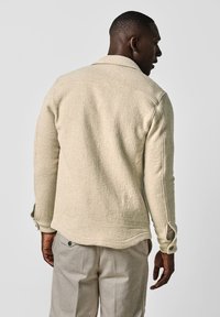 Camicia beige a maniche lunghe con texture, colletto, bottoni ai polsini e orlo dritto, indossata sopra pantaloni grigio chiaro, vista da dietro.