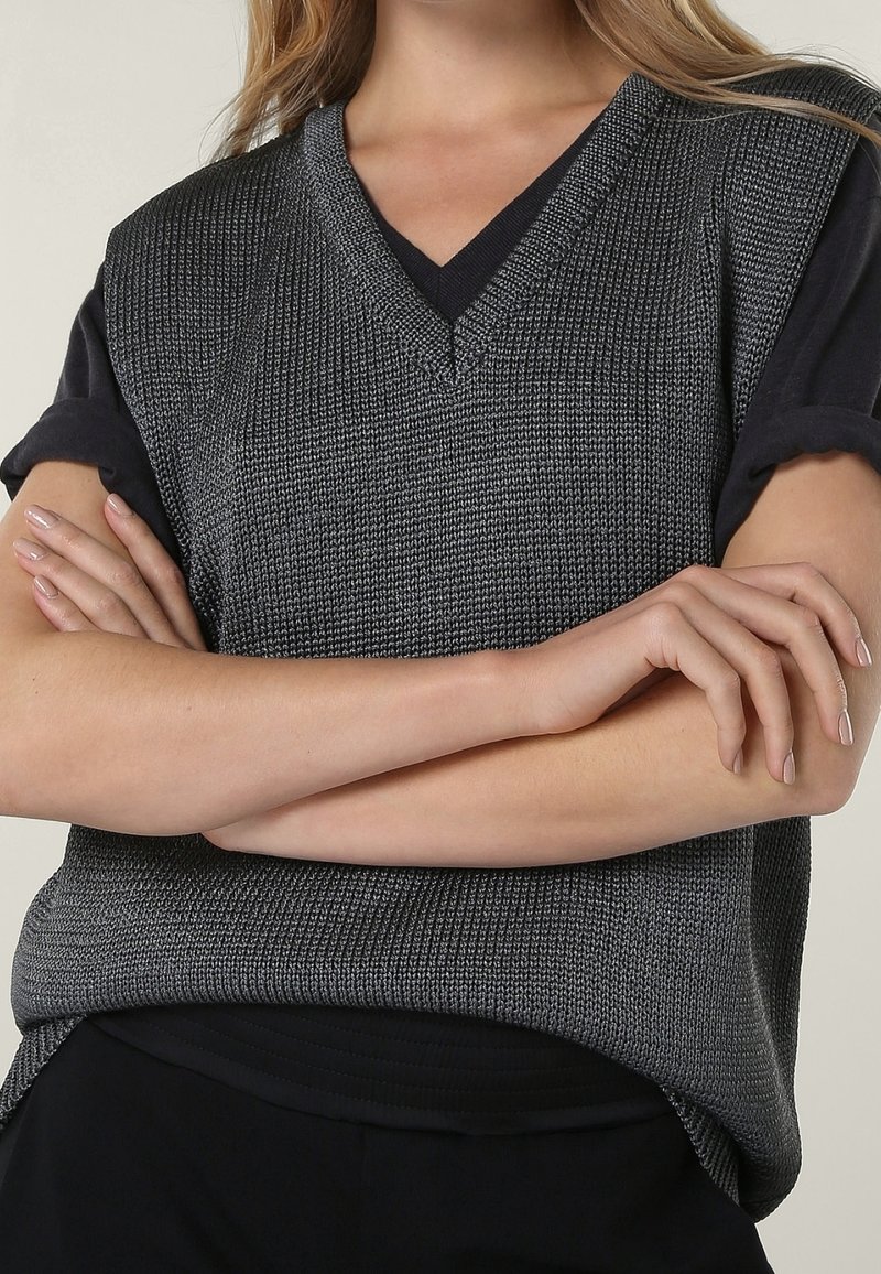 Gilet gris tricoté avec un col en V, présentant un tissu texturé et des manches courtes noires. Les bras du modèle sont croisés devant.
