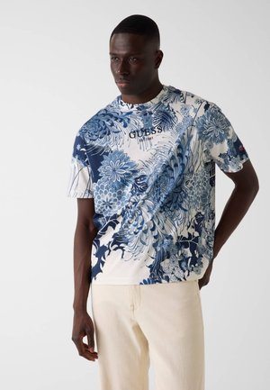 Mężczyzna w krótkim rękawie koszulce z niebieskim kwiatowym wzorem i napisem "GUESS est. 1981", w połączeniu z jasnobeżowymi spodniami.