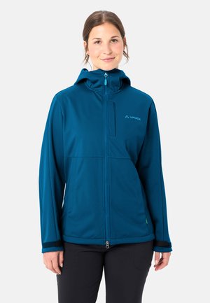 Donna che indossa una giacca con cappuccio blu verde con zip e logo sulla tasca sul petto, in piedi davanti a uno sfondo chiaro e semplice.