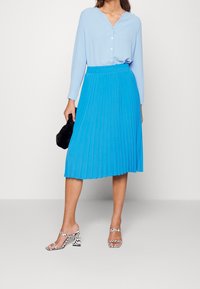 Blusa azul claro con escote en V y botones, combinada con una falda midi plisada azul brillante y sandalias de tacón cuadrado de diseño. Clutch negro.