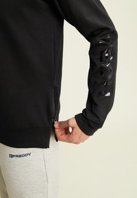 Persona che indossa un felpa nera con il testo "FREDDY" sulla manica, mentre tira la zip sul lato; i pantaloni grigi chiari mostrano anch'essi il logo "FREDDY".