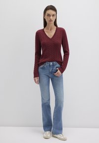 Bordeauxfarbener Grobstrickpullover mit V-Ausschnitt, kombiniert mit hellblauen Schlagjeans. Das Modell trägt beigefarbene Sneakers. Schlichtes, modernes Design.