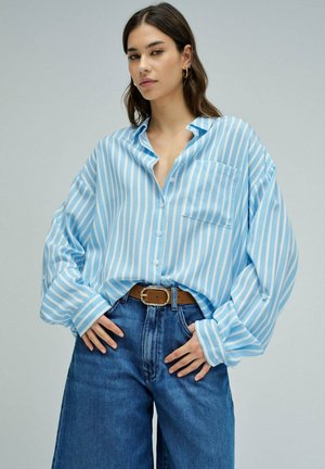 Frau trägt ein übergroßes hellblau-weiß gestreiftes Hemd, in hoch taillierte blaue Jeans mit braunem Gürtel gesteckt, steht mit den Händen in der Nähe der Taille.
