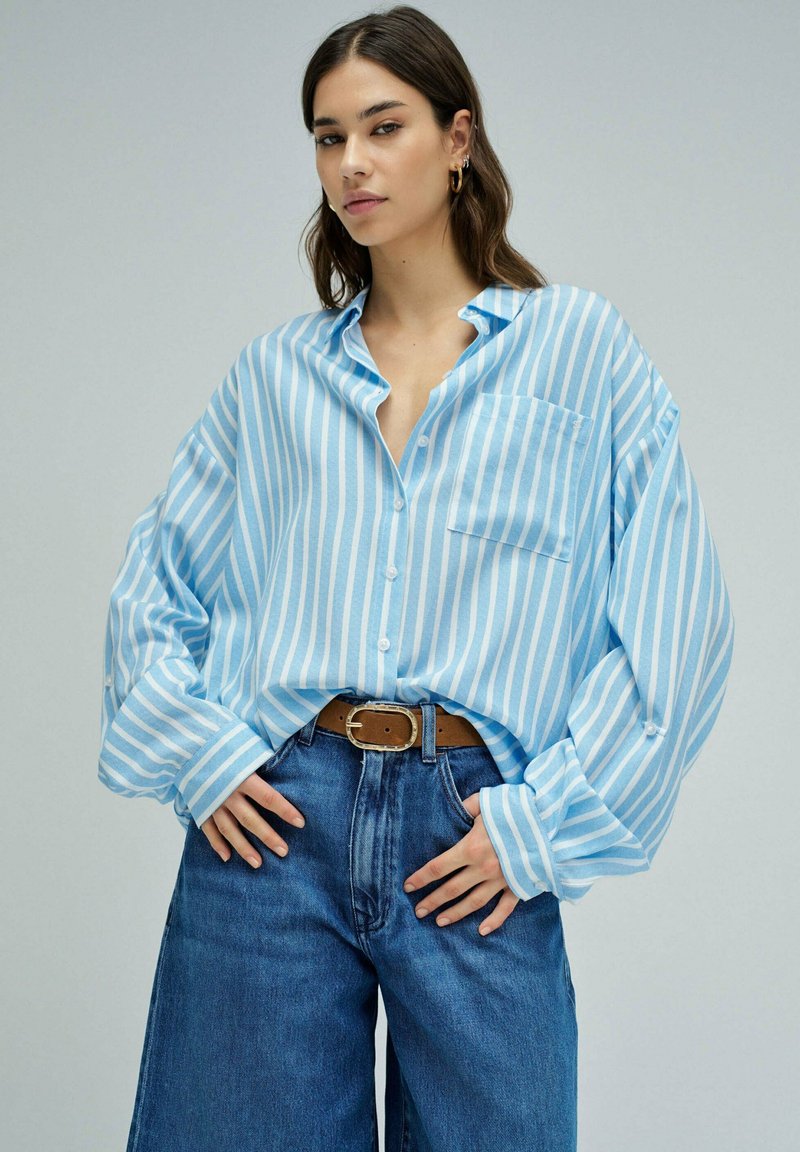 Woman wearing oversized light blue and white striped shirt tucked into high-waisted blue jeans with brown belt, standing with hands near waist.
