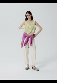 Žena s dlhými tmavými vlasmi, oblečená v žltom kostkovanom topu bez rukávov, bielych nohaviciach, strieborných balerínach a fialovom svetri zviazanom okolo pása.