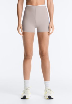 Femme portant un short de sport beige près du corps, des chaussettes blanches et des baskets blanches avec des accents jaunes, debout sur un fond blanc uni.
