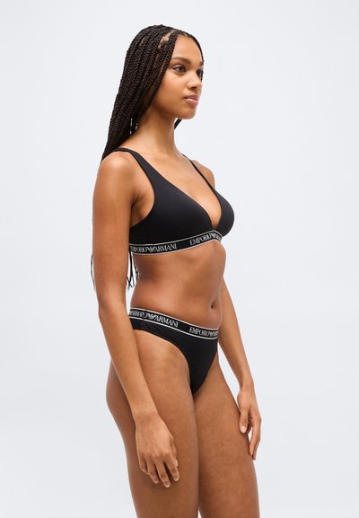 Γυναίκα με μακριά πλεξούδες που φοράει μαύρο σουτιέν και εσώρουχα Emporio Armani, στέκεται πλαγίως μπροστά σε ένα απλό ανοιχτό φόντο.