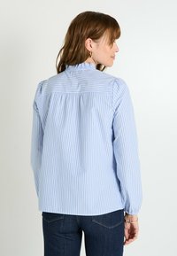 Blusa de rayas azul claro y blanca con un escote con volantes, mangas largas y panel trasero fruncido. Hecha de una tela suave con un ligero brillo.