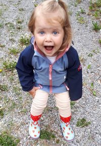 Enfant portant une veste bleue et marine avec des accents rose, un pantalon crème, et des bottes de pluie rouges et bleues ornées de motifs de fraises.