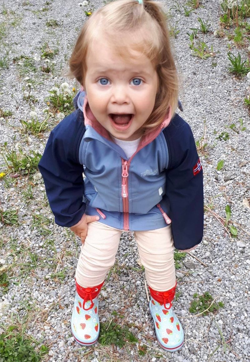 Enfant portant une veste bleue et marine avec des accents rose, un pantalon crème, et des bottes de pluie rouges et bleues ornées de motifs de fraises.